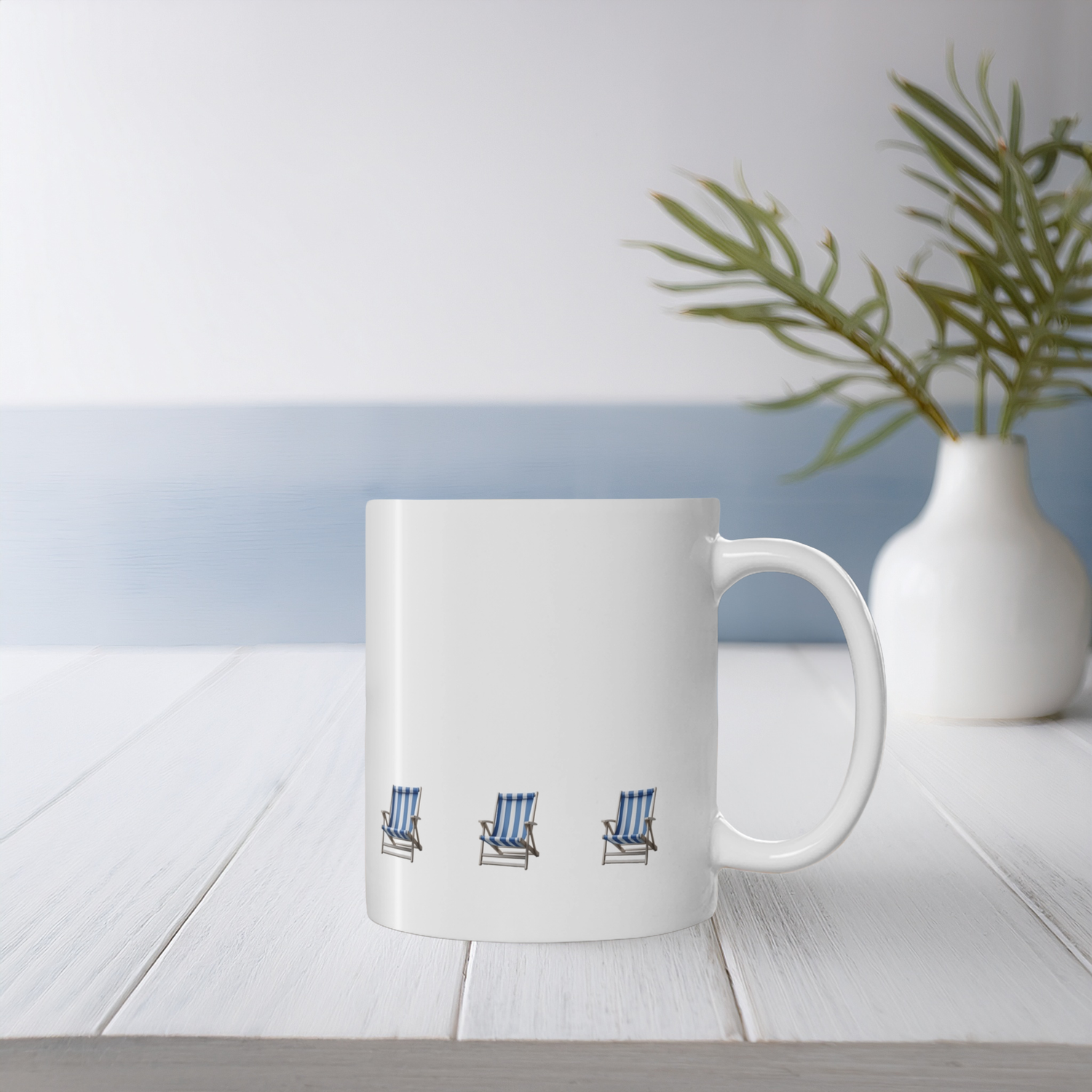 White ceramic mug with blue and white stripe beach chair design on a white wooden table with small vase & wildflowers. The beach chairs are in a row near the bottom of the mug.