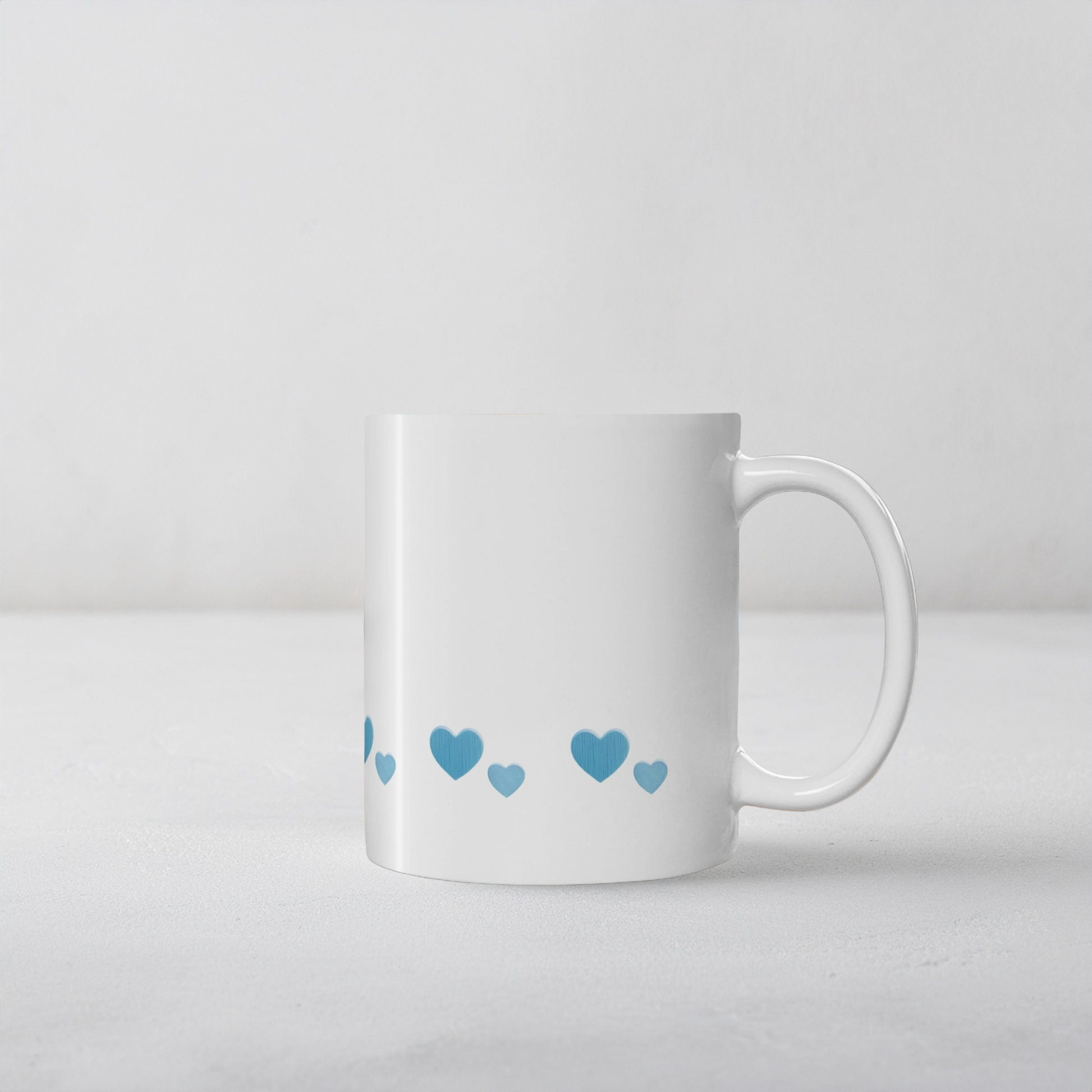 White ceramic mug with blue heart design on a white cement style surface. The blue hearts are in a row near the bottom of the mug.