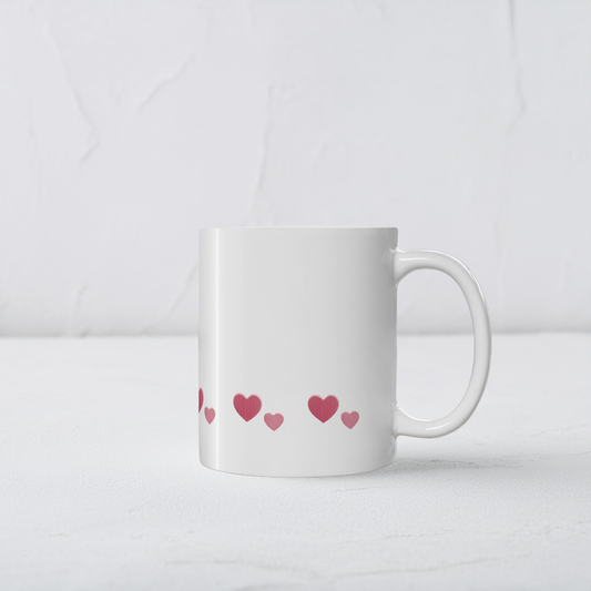 White ceramic mug with row of red hearts wrapping around the mug near the bottom. The mug sits on a white wooden surface