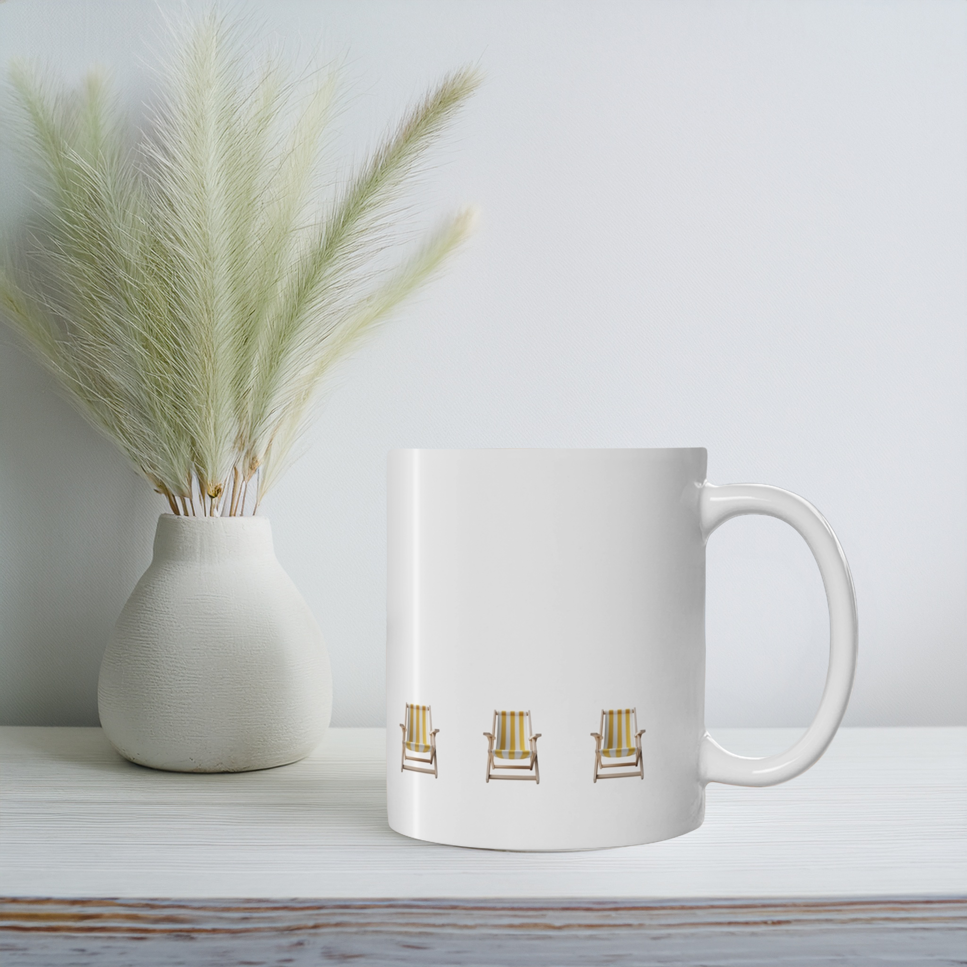 White ceramic mug with yellow and white stripe beach chair design on a white wooden shelf with small vase & wildflowers. The beach chairs are in a row near the bottom of the mug.