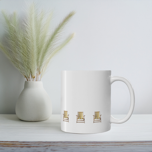 White ceramic mug with yellow and white stripe beach chair design on a white wooden shelf with small vase & wildflowers. The beach chairs are in a row near the bottom of the mug.