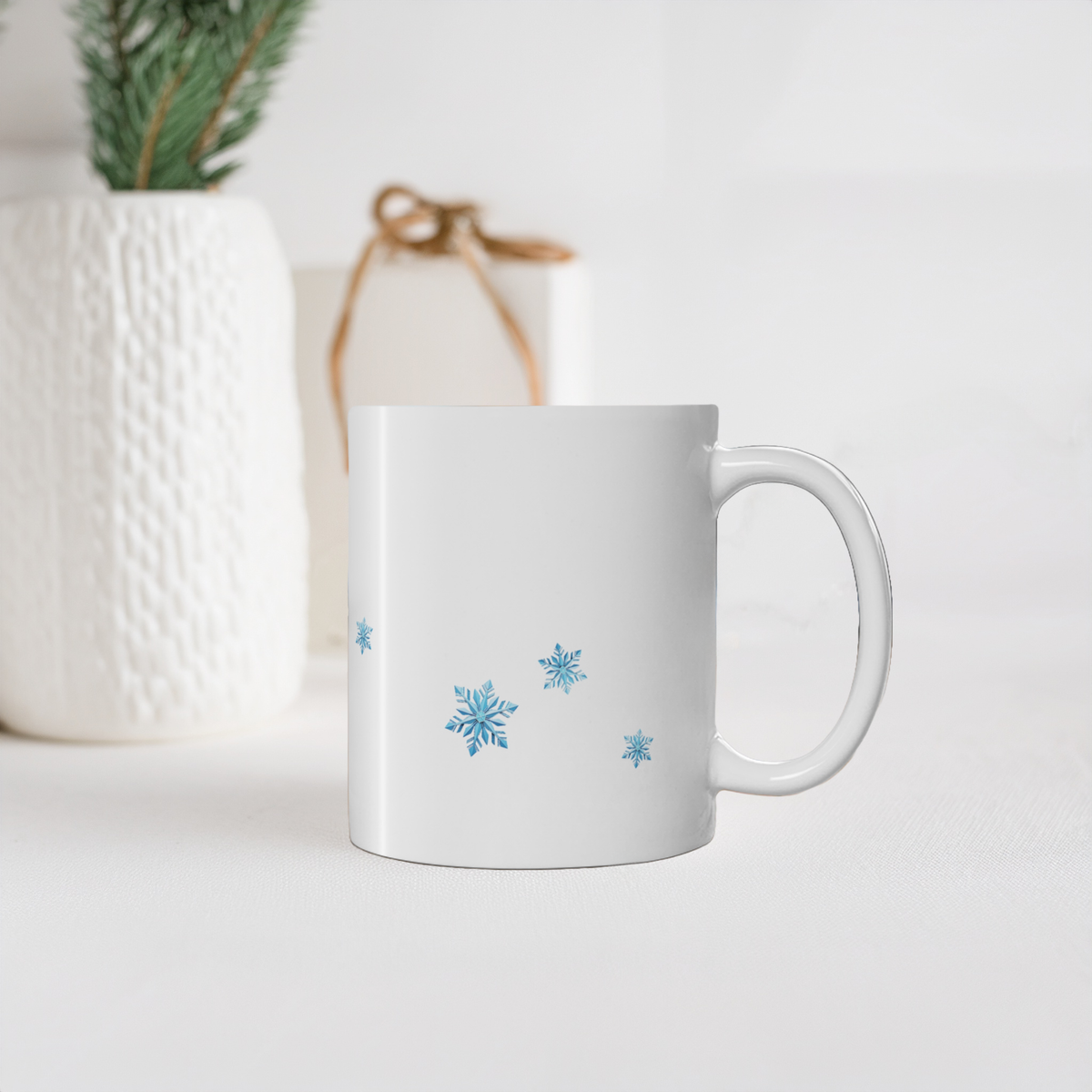 A white ceramic mug decorated with blue snowflakes, set against a softly blurred background with a white textured vase and a small wrapped gift.