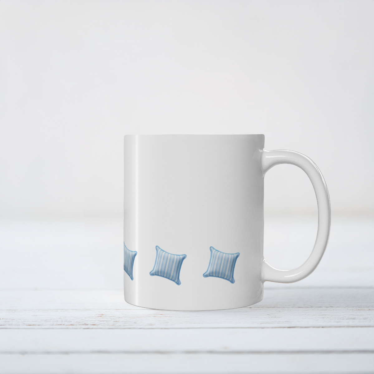 A white ceramic coffee mug with a handle, featuring a printed pattern of three blue striped pillows near the bottom, set against a white wooden background.