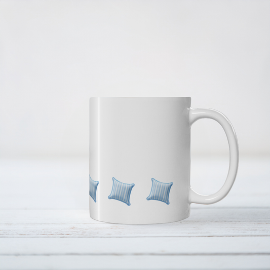 A white ceramic coffee mug with a handle, featuring a printed pattern of three blue striped pillows near the bottom, set against a white wooden background.