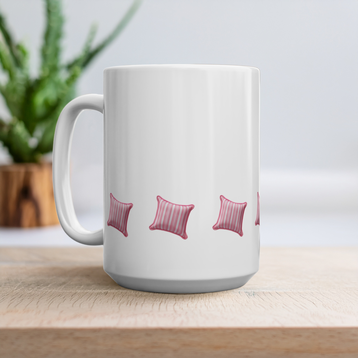 15oz white ceramic mug with a handle, decorated with a repeating pattern of pink and white striped square pillows, sitting on a light wooden surface with a blurred green plant in the background.