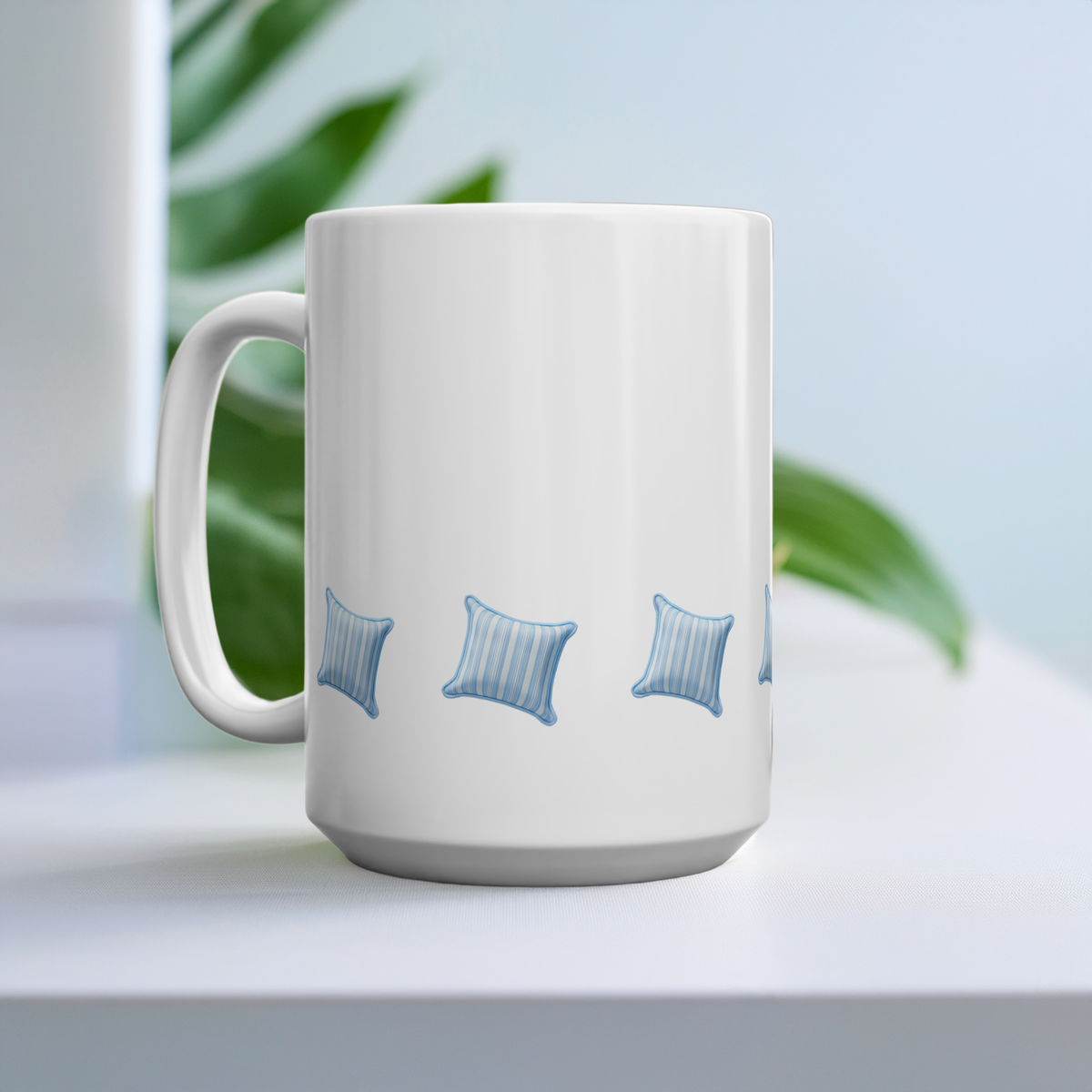 A 15oz white ceramic mug with a handle, decorated with a repeating pattern of small blue striped pillows, set on a white surface with a soft-focus background of green leaves and a light blue wall.