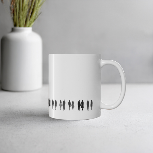 White ceramic mug with black line art illustration of people holding hands. The mug is set on a light grey surface with a blurred white vase containing dried grass in the background.