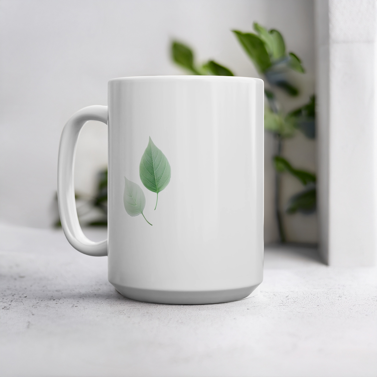 White ceramic 15oz mug with two green and white autumn leaf design. The mug sits on cement style surface with plant in the background.