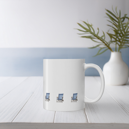 White ceramic mug with blue and white stripe beach chair design on a white wooden table with small vase & wildflowers. The beach chairs are in a row near the bottom of the mug.