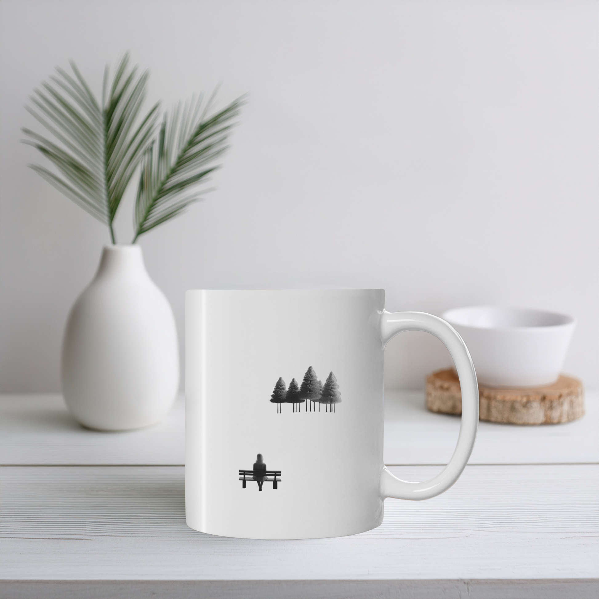 White ceramic mug with black line art illustration of woman on bench looking at trees. The mug sits on a white wooden surface with small plant pot and ceramic ornament. 