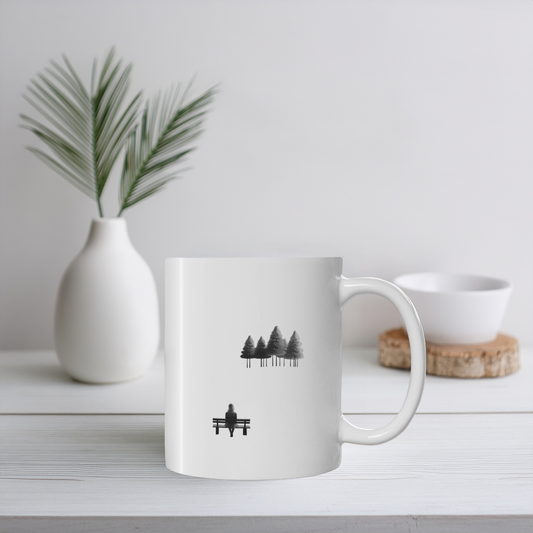 White ceramic mug with black line art illustration of woman on bench looking at trees. The mug sits on a white wooden surface with small plant pot and ceramic ornament. 