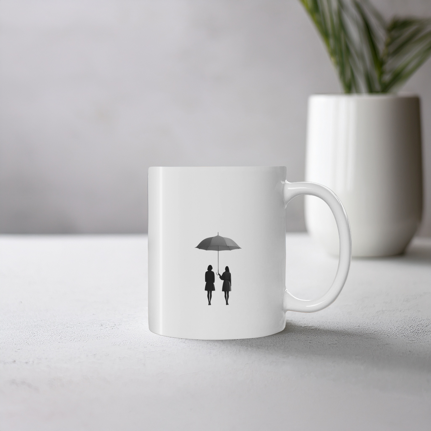 White ceramic mug with black line art illustration of two friends under an umbrella. The mug sits on a cement style surface with small plant pot behind it.